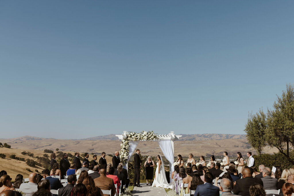 Overview of wedding ceremony at Willow Heights mansion with white wedding arch and flowers with rolling hills in background