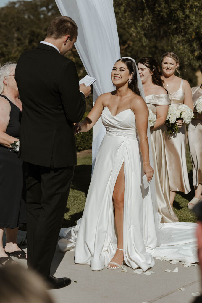 Bride smiling while holding groom's hand as he reads vows during wedding ceremony with bridesmaids smiling in background at Willow Heights mansion