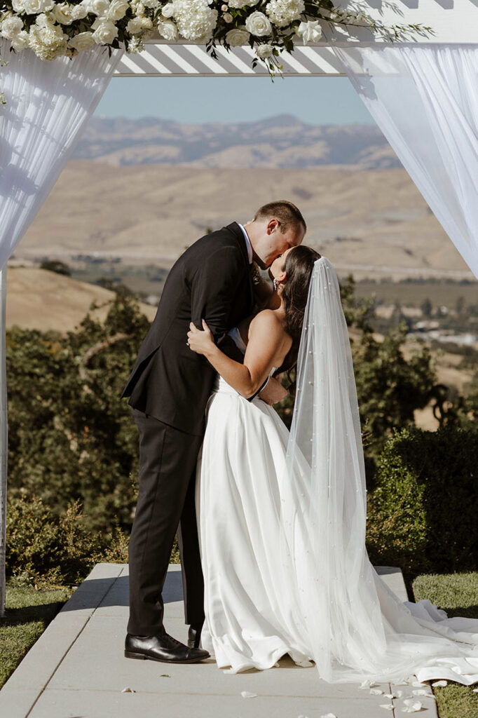 Wedding couple kissing under white wedding arch during ceremony at Willow Heights mansion with rolling hills in background