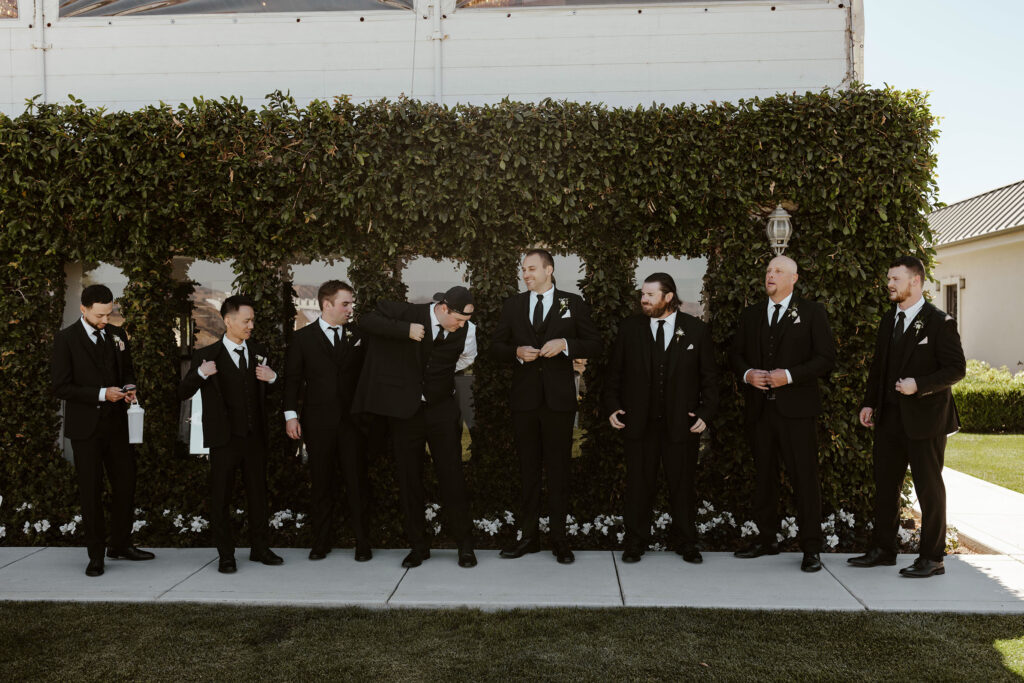 Wedding groom and groomsmen buttoning and putting on jackets while in front of grass hedge with windows at Willow Heights mansion