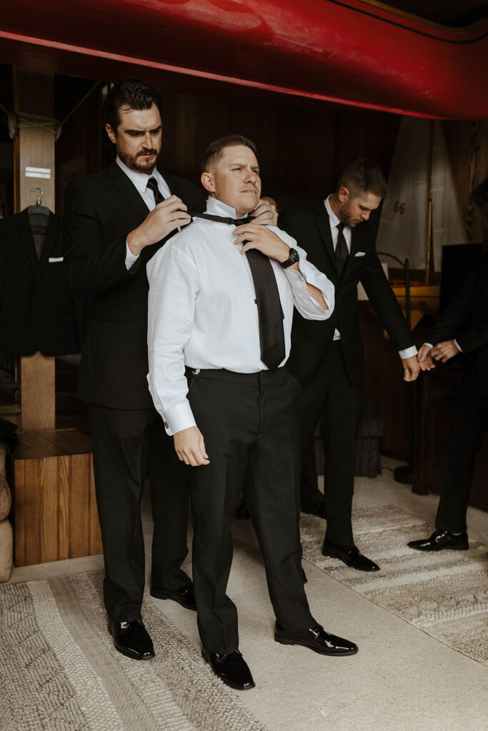 Wedding groom holding tie as groomsmen helps adjust collar behind him while inside at the PlumpJack Inn