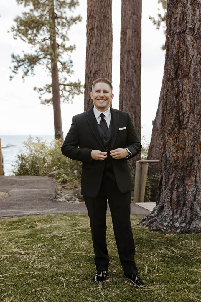 Wedding groom buttoning suit jacket while standing on grass outside of the PlumpJack Inn with Lake Tahoe in background