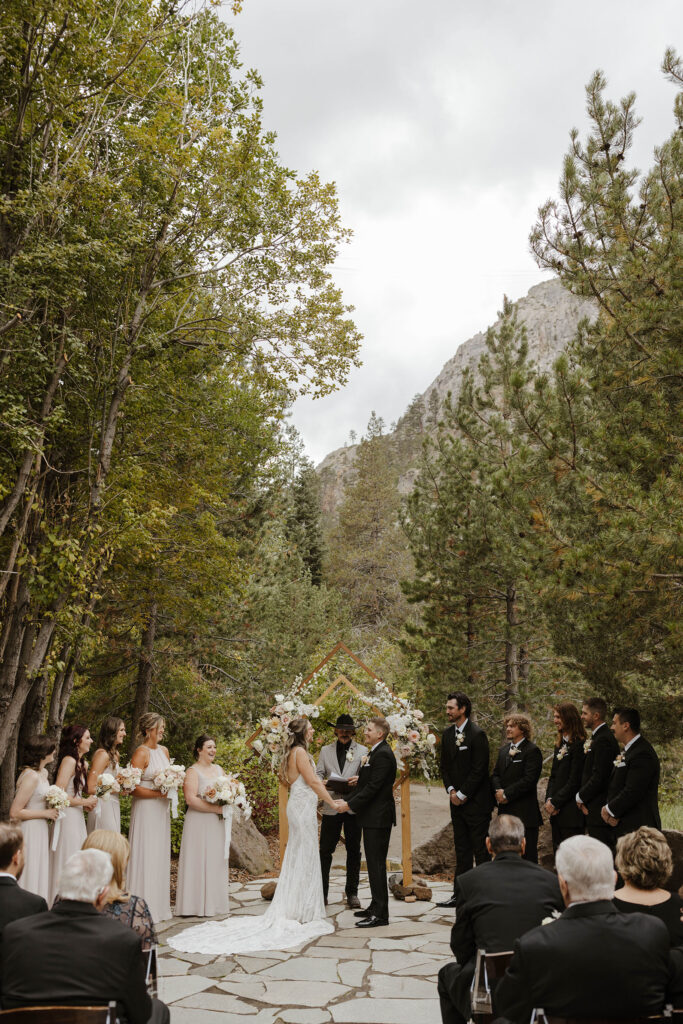Wedding ceremony at the PlumpJack Inn with couple holding hands under the arch with bridal party on either side and pine trees in background