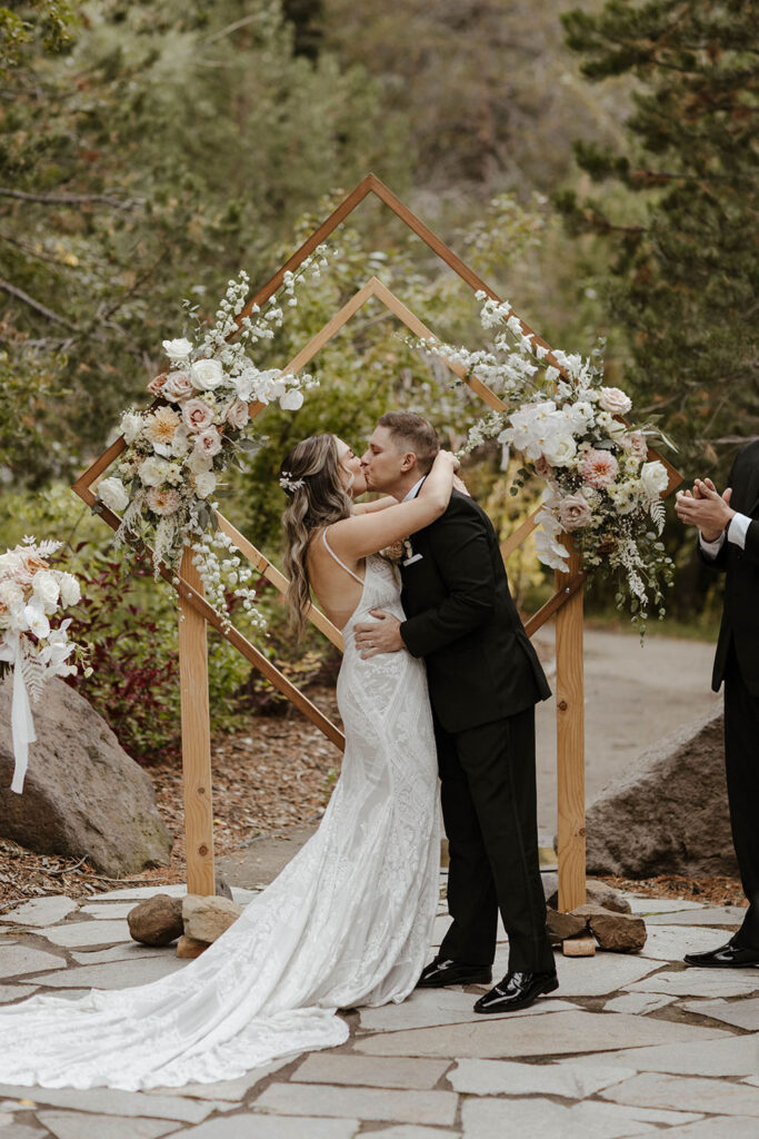 Wedding couple kissing in front of wedding arch during fall ceremony at the PlumpJack Inn with pine trees in background