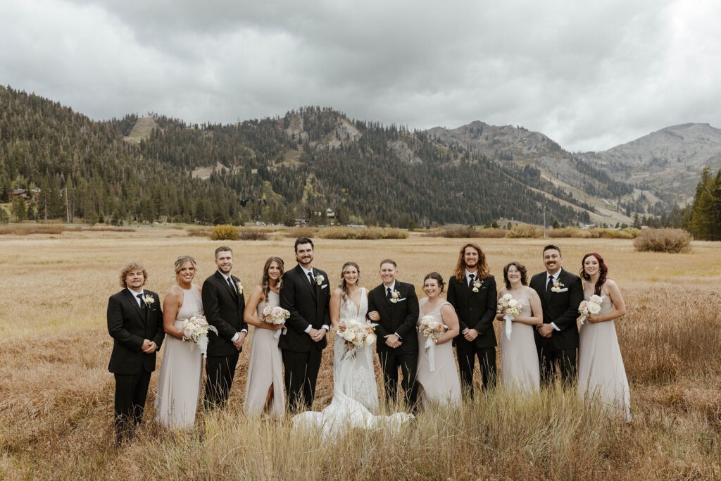 Wedding couple and bridal party standing next to each other and smiling at camera while in golden field at the PlumpJack Inn with mountains in background