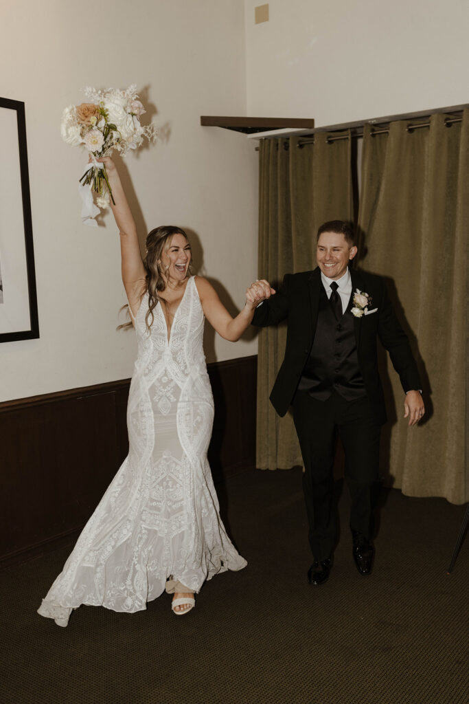 Wedding couple holding hands and smiling while bride holds floral bouquet up during grand entrance at the PlumpJack Inn