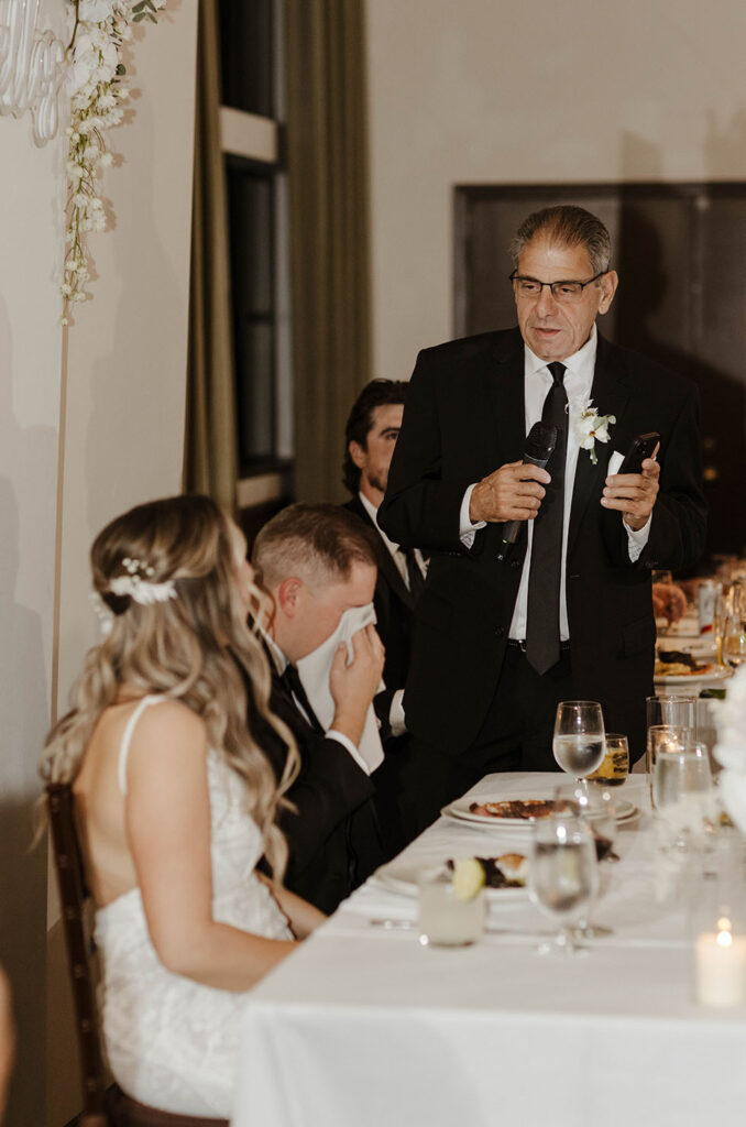 man giving a wedding speech while the groom cries into a napkin