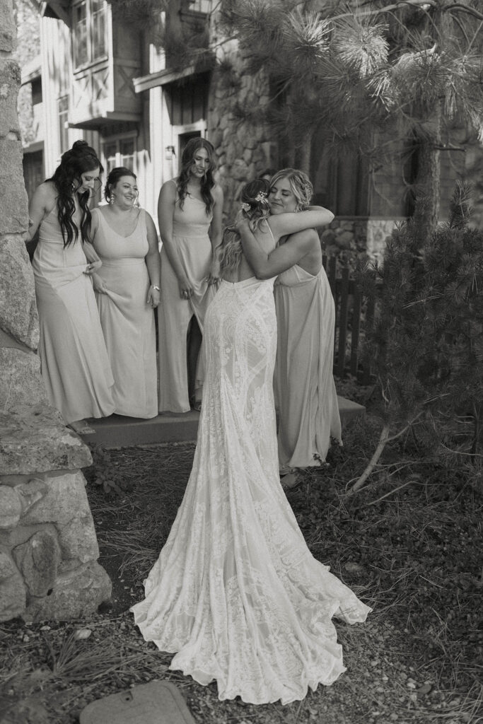 Wedding bride hugging bridesmaids after first look with dress outside at the PlumpJack Inn with wooden buildings in background