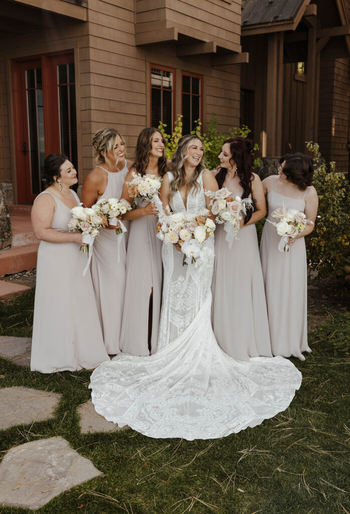 Wedding bride holding floral bouquet while standing next to bridesmaids while smiling at the PlumpJack Inn with wooden buildings in background