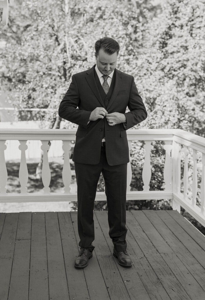 Wedding groom buttoning suit jacket while standing on wooden patio at Logan Shoals