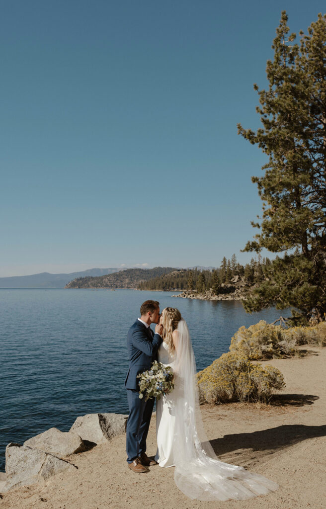 Wedding groom kissing bride while holding her chin and she holds floral bouquet on sandy trail next to Lake Tahoe at Logan Shoals
