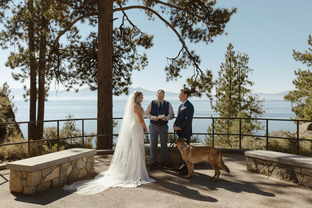 Wedding couple smiling at each other while groom holds dog's leash during ceremony at Logan Shoals with Lake Tahoe and trees in background