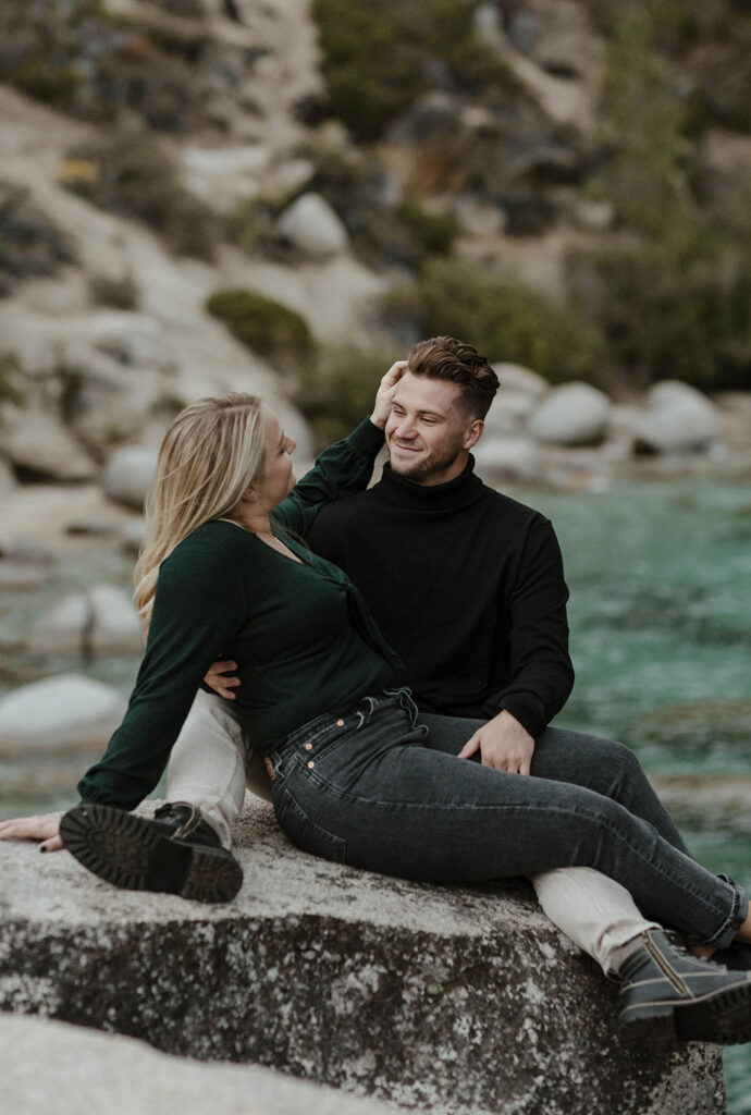 Woman playing with fiancé's hair while smiling at each other and sitting on rock together at Lake Tahoe