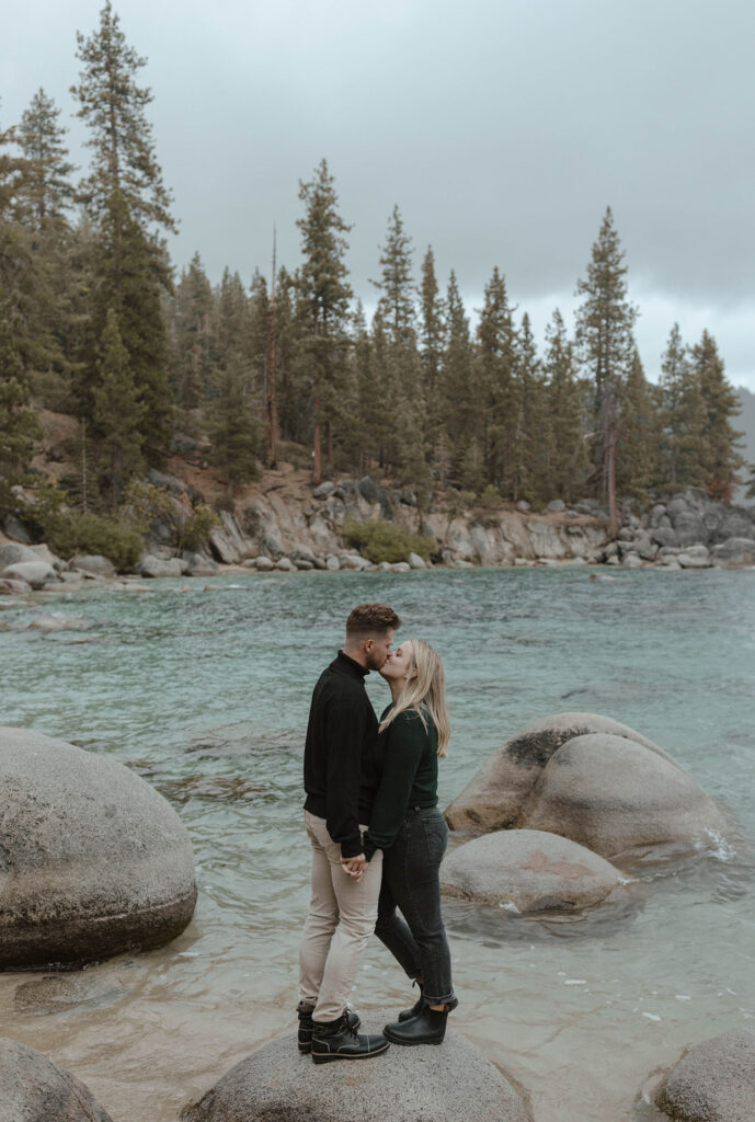 Engagement couple holding hands while standing on a rock in water while kissing at Lake Tahoe with pine trees and lake in background