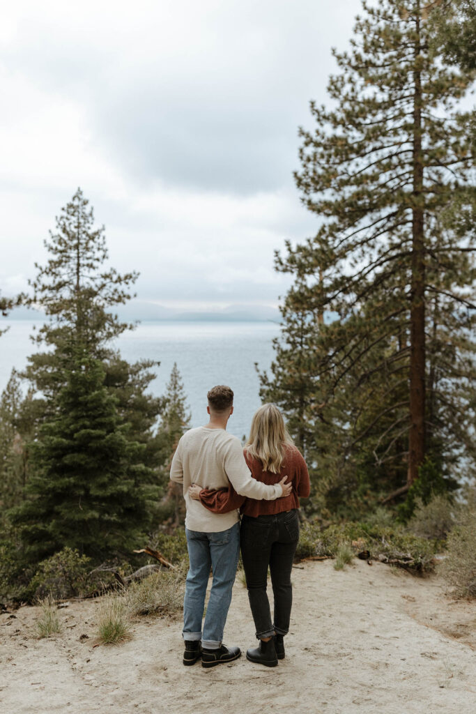 Engagement couple with arms around each other facing away from camera and looking out over Lake Tahoe in between pine trees