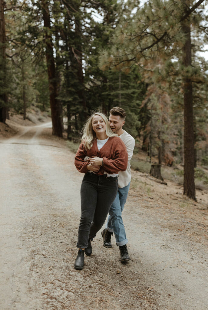 Woman smiling at camera while fiancé hugs her from behind while laughing and running along dirt trail together in Lake Tahoe