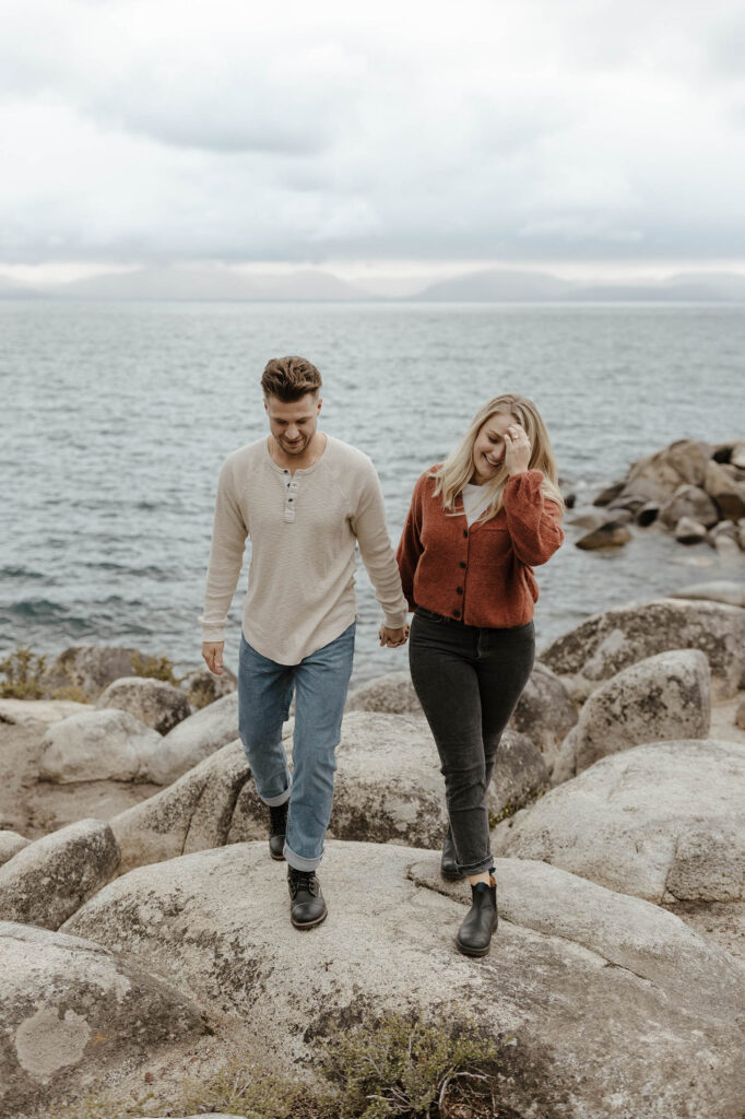 Engagement couple holding hands and walking across large rocks together with Lake Tahoe in background