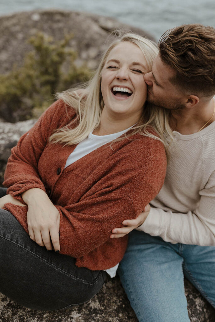 Woman laughing while fiancé hugs her and kisses her cheek while both sit on rock in Lake Tahoe