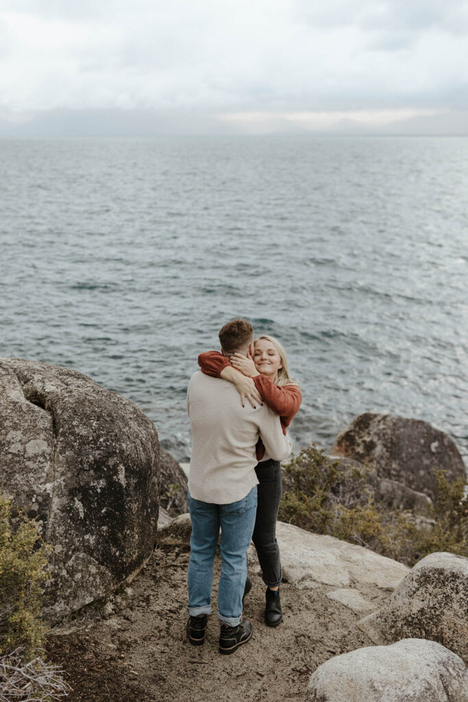 Engagement couple hugging and smiling while standing on large rock with Lake Tahoe in background