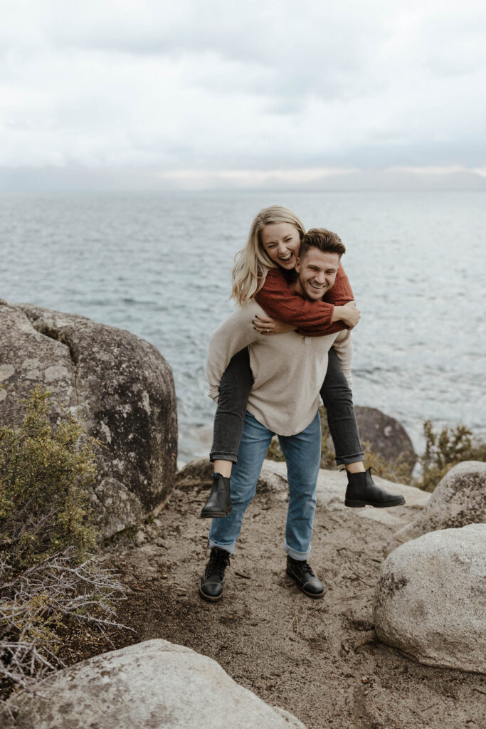 Man giving fiancé a piggyback ride while both laugh and stand on large rock with Lake Tahoe in background