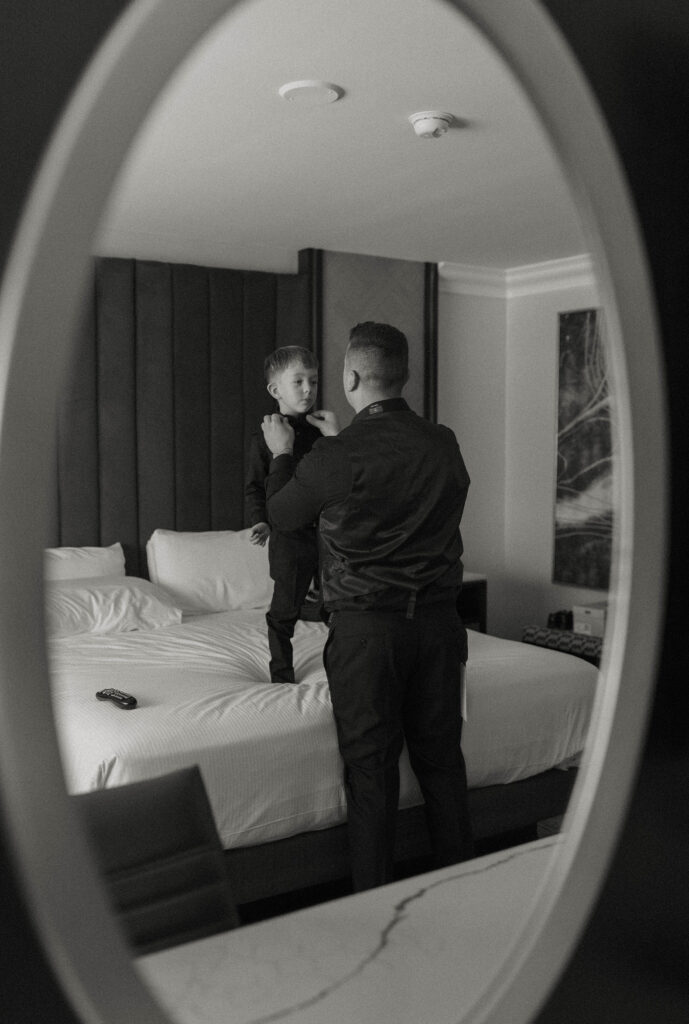 Wedding groom adjusting bowtie on son standing on hotel room bed reflection in a mirror at Sierra Water Gardens