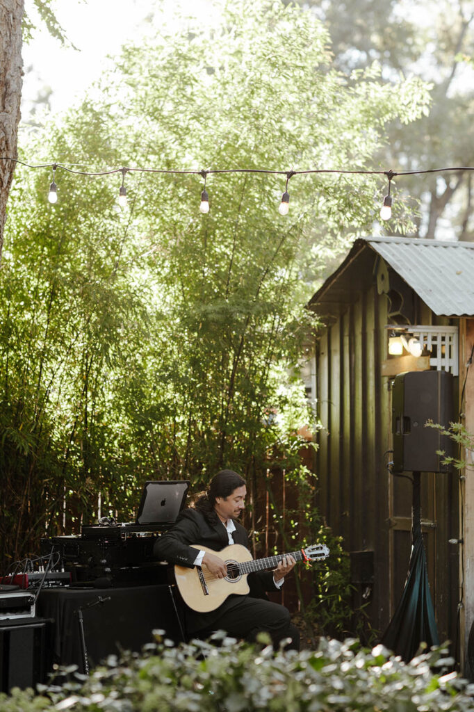 Wedding dj playing guitar in front of audio equipment surrounded by green florals at Sierra Water Gardens