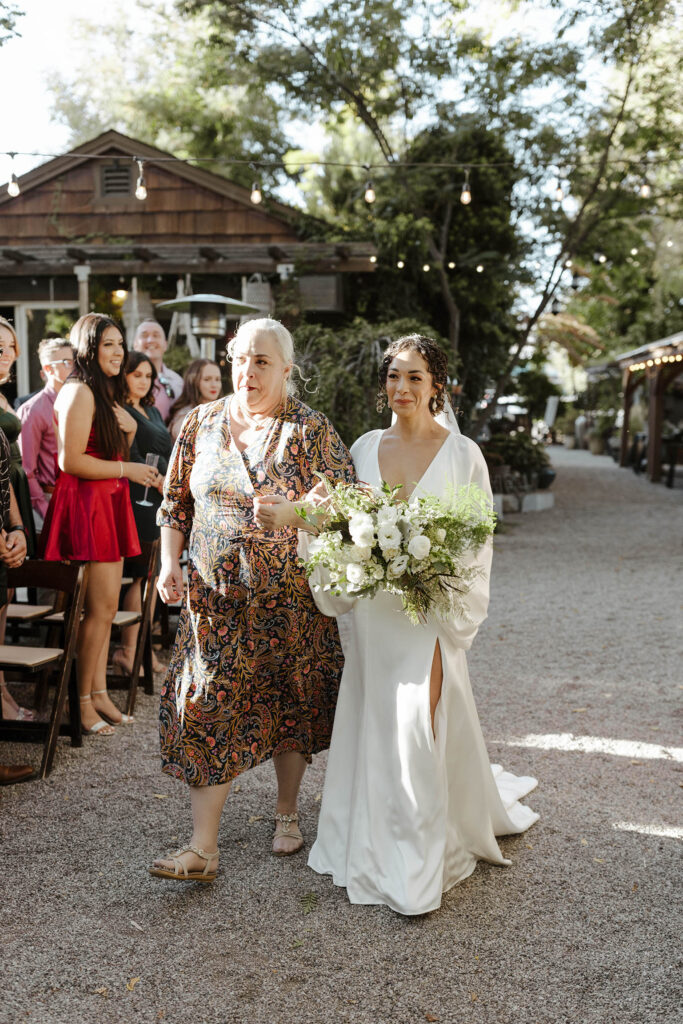 Bride walking down wedding aisle while holding mom's arm and white floral bouquet at Sierra Water Gardens