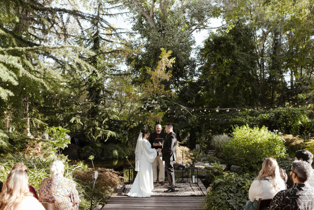 Wedding couple holding hands during ceremony at Sierra Water Gardens with string lights and lots of greenery in background