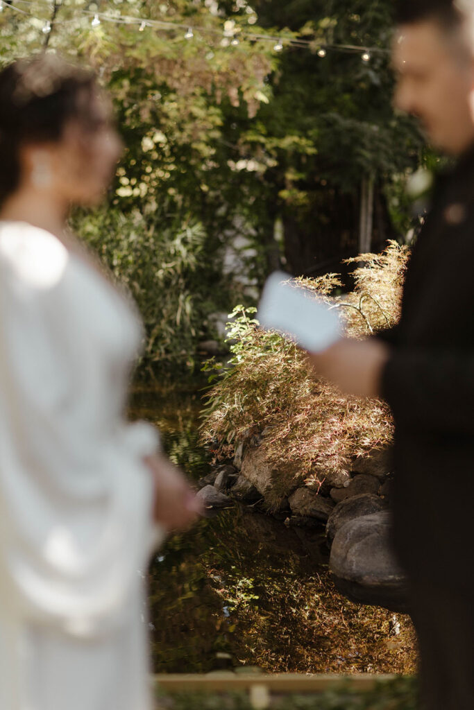 Wedding groom reading vows to bride while camera focuses on background at Sierra Water Gardens