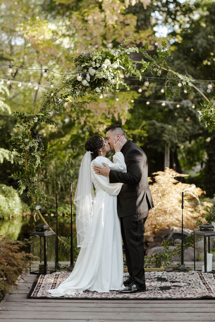 Wedding couple kissing while holding each other after ceremony with wedding arch and lots of greenery at Sierra Water Gardens