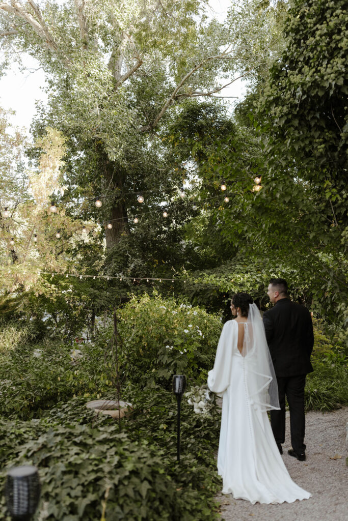 Wedding couple holding hands while walking away from camera through garden with lots of greenery and string lights at Sierra Water Gardens