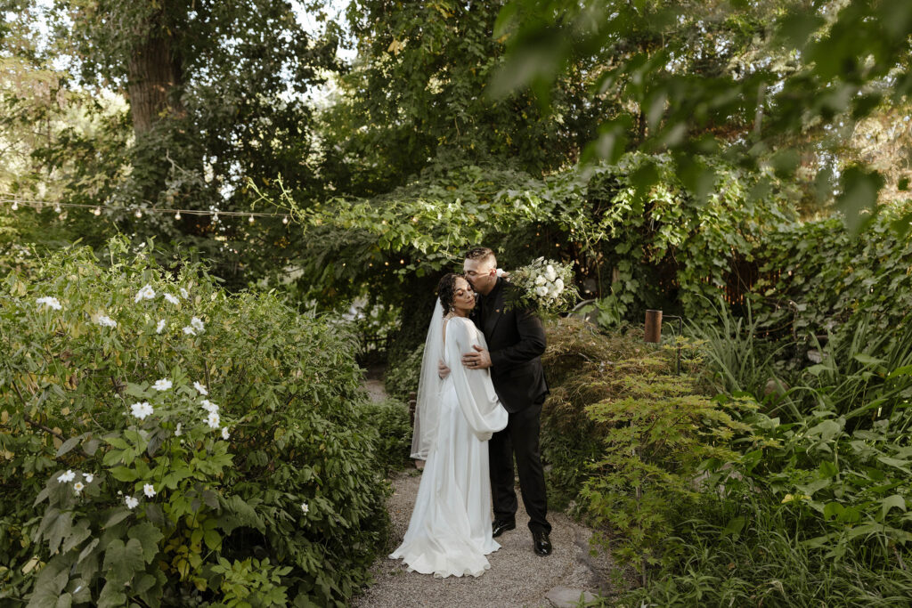 Wedding couple standing in garden surrounded by greenery while groom kisses bride on the cheek at Sierra 
Water Gardens