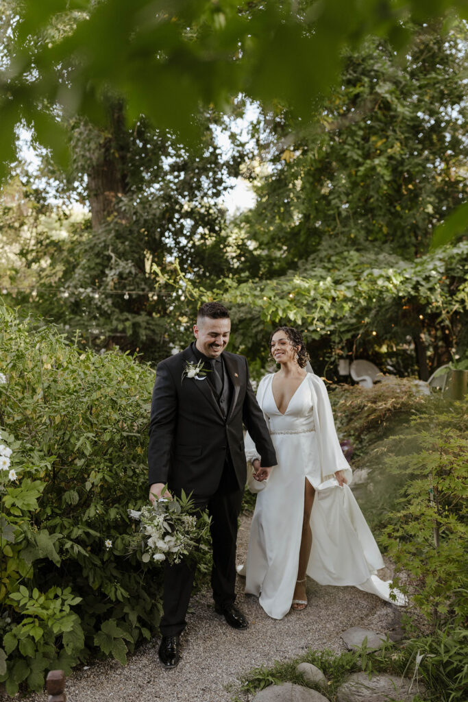 Wedding couple holding hands while walking through lots of greenery and smiling at the Sierra Water Gardens