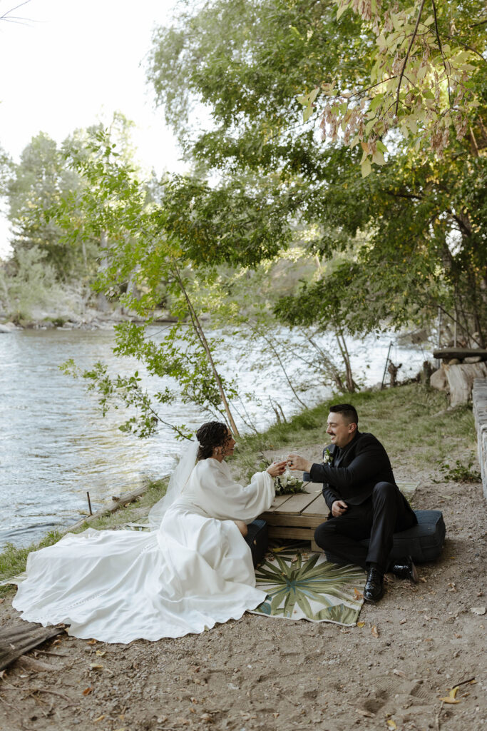 Wedding couple sitting by table next to the water while cheering drink glasses with trees in background at Sierra Water Gardens