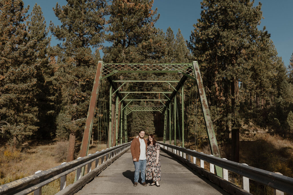 Engagement couple with arms around each other looking at camera while standing in front of bridge at Graeagle