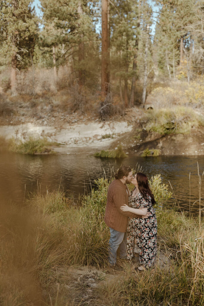 Picture through the bushes of engagement couple holding each other and kissing while standing next to river at Graeagle