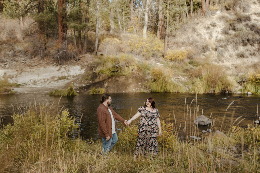 Engagement couple holding hands while woman looks back at fiancé walking next to river at Graeagle