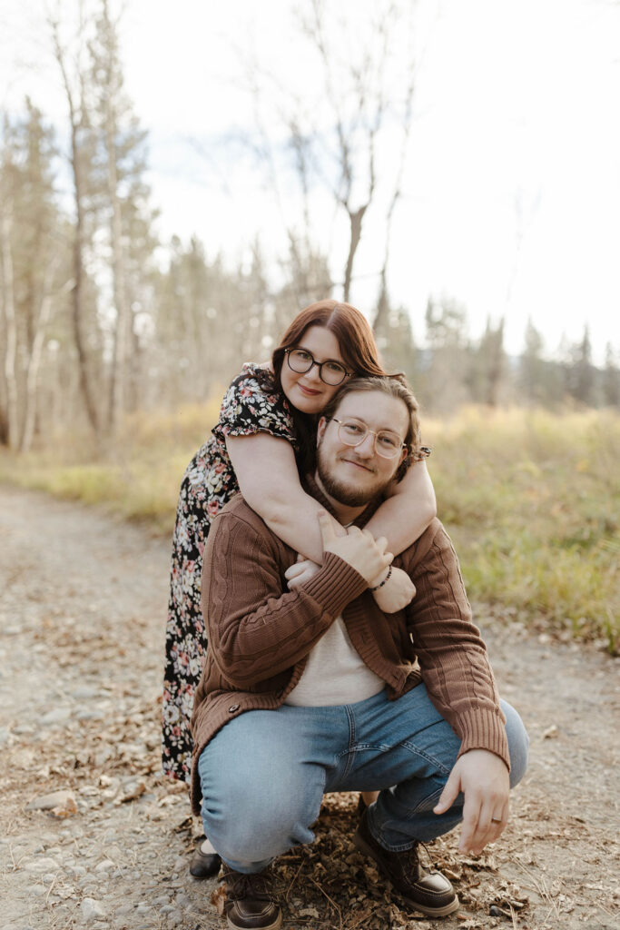 Woman hugging fiancé from behind while they both crouch and smile at the camera at Graeagle