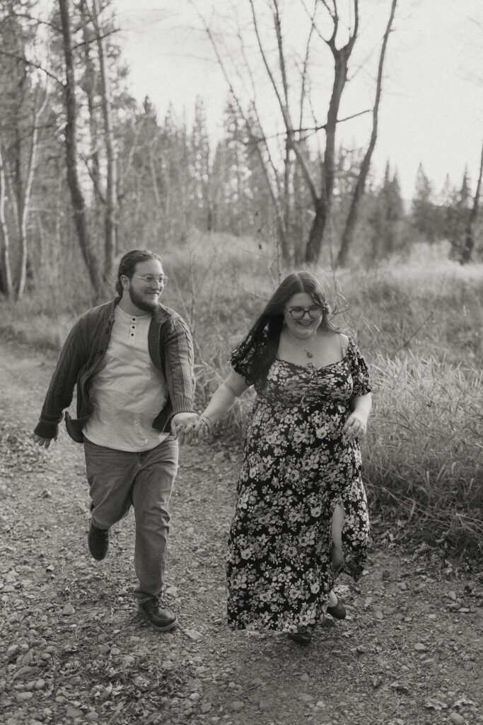 Engagement couple running down dirt trail together while smiling and holding hands with trees in background at Graeagle