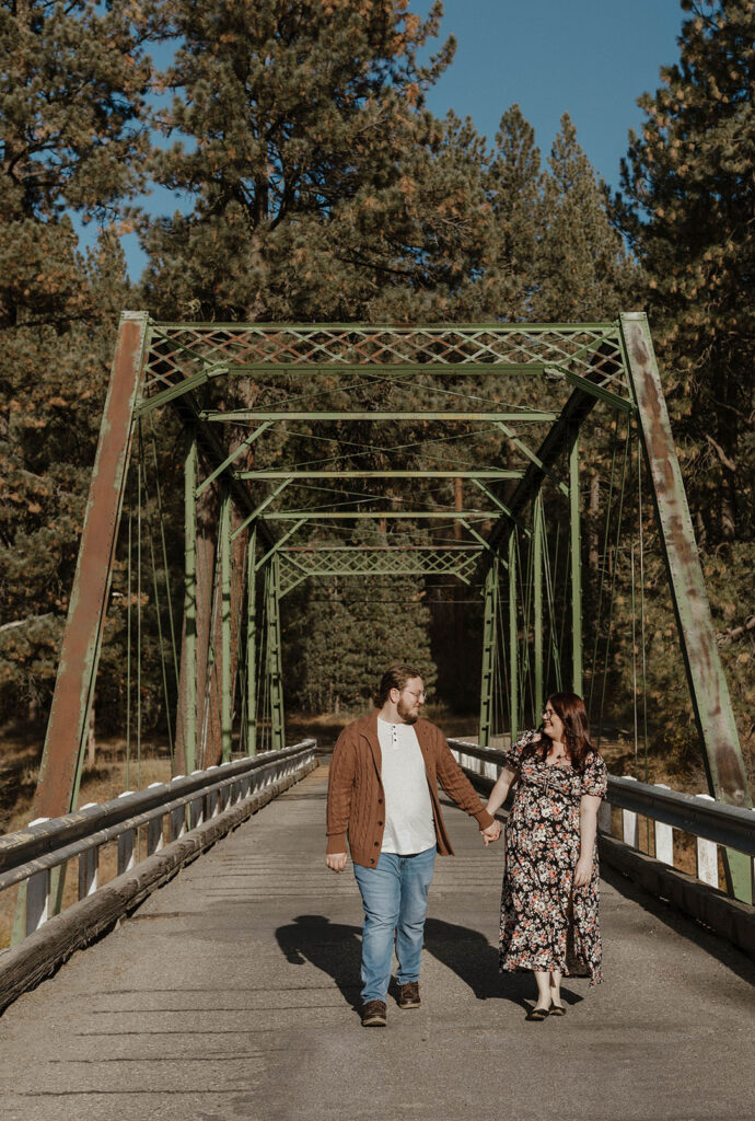 Engagement couple holding hands while walking under bridge and smiling at each other at Graeagle