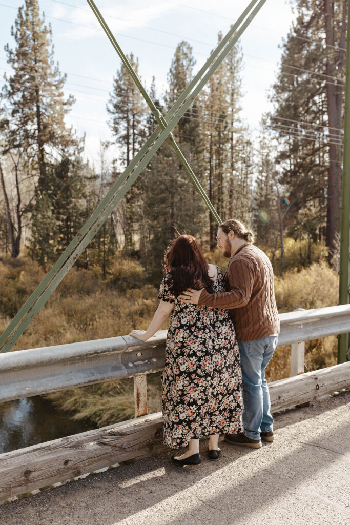 Man with hand on fiancé's back while they stand at the edge of a bridge and look at the river together at Graeagle