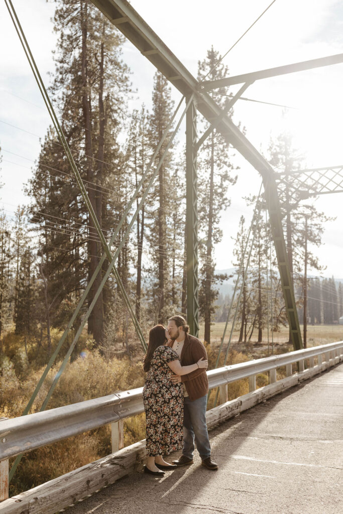Engagement couple holding each other while kissing on a bridge in Graeagle with trees and field in background