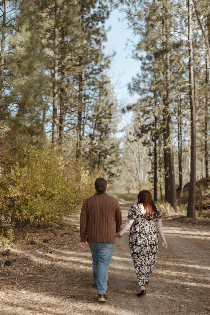 Engagement couple holding hands while walking away from camera down dirt trail together with tall pine trees on either side at Graeagle