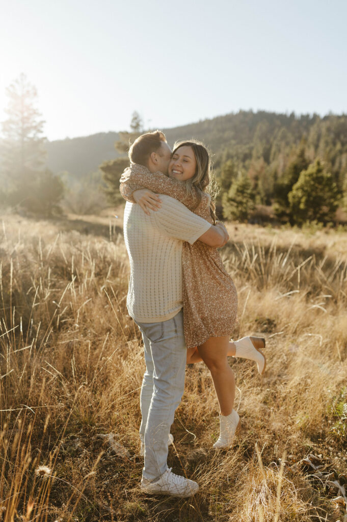 Engagement couple hugging while in golden field and woman smiles in Lake Tahoe with trees in background