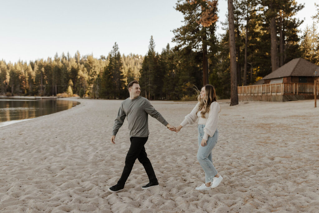 Engagement couple holding hands while walking along sandy beach together and man looks back over shoulder at fiancé in Lake Tahoe