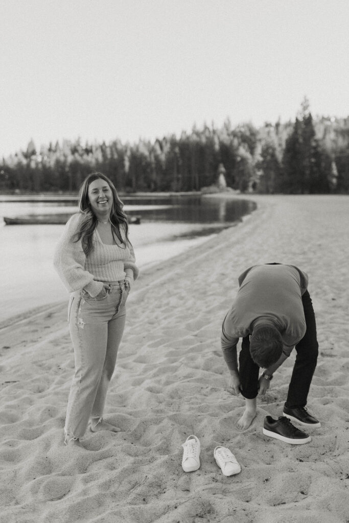 Man taking shoes and socks off while woman smiles at camera with hands in pocket while on a sandy beach in Lake Tahoe