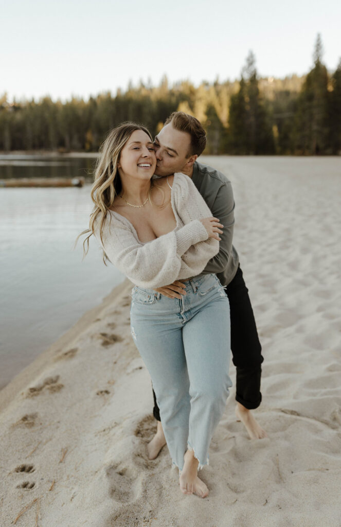  Man hugging fiancé from behind while she smiles while on a sandy beach in Lake Tahoe with water in background
