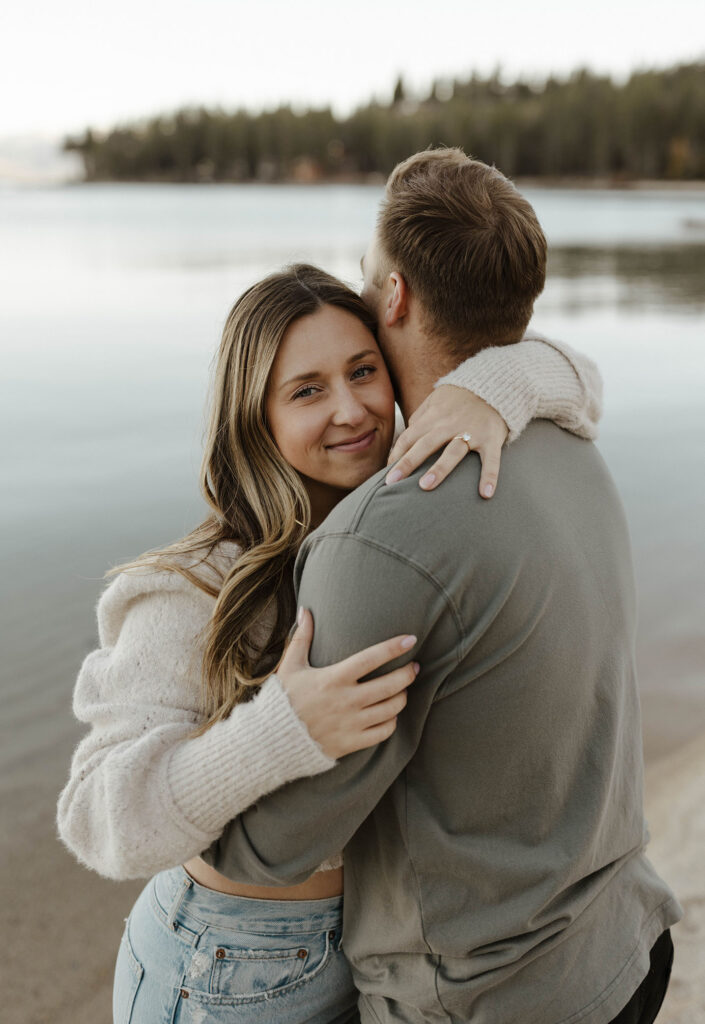 Woman smiling at camera while hugging fiancé at Lake Tahoe with water in background