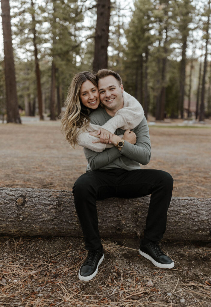 Engagement couple sitting on log together smiling at camera at Lake Tahoe with tall trees in background