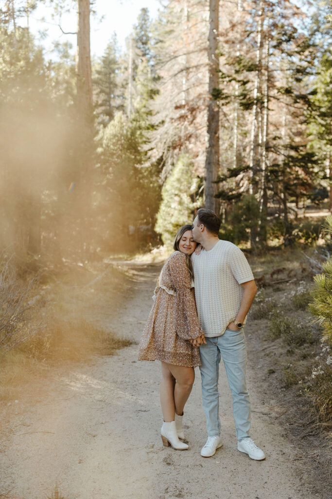 Man kissing fiancé on forehead while both stand together on trail holding hands in Lake Tahoe with trees in background